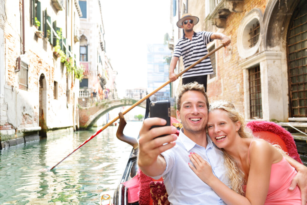 Couple in Venice on Gondole ride romance in boat happy together on travel vacation holidays. Romantic young beautiful couple taking self-portrait sailing in venetian canal in gondola. Italy. Romantic getaway. Allegro Luxury Vacations. Brenda Ajay, Virtuoso luxury travel advisor.