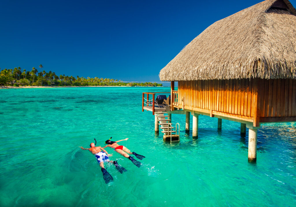 Young couple snorkeling from hut over blue tropical lagoon. Allegro Luxury Vacations romantic getaway. Brenda Ajay, Virtuoso luxury travel advisor.