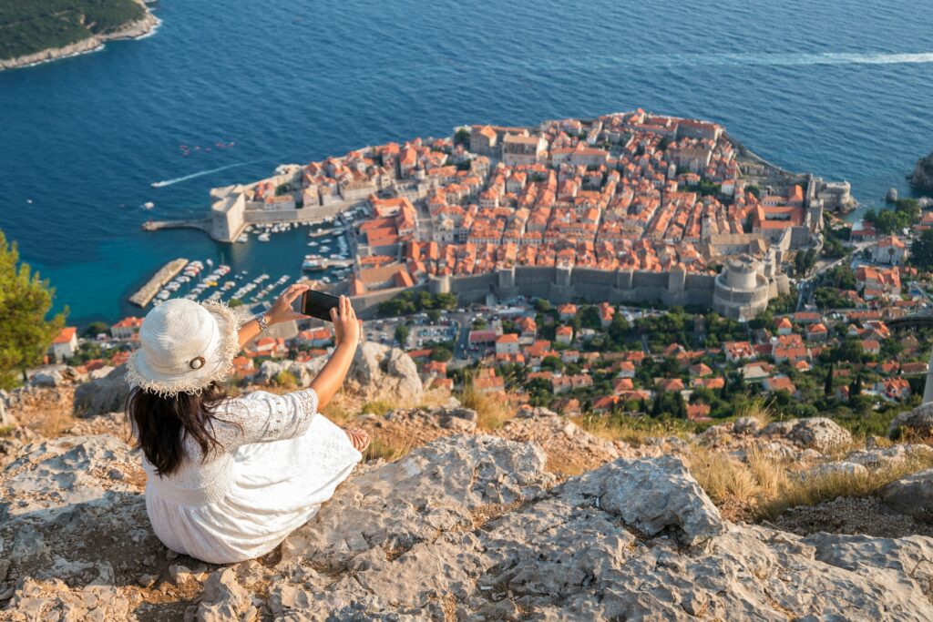 A beautiful woman sitting on rocks taking a photo of Dubrovnik Old Town below. Allegro Luxury Vacations. Romantic getaway. Brenda Ajay, travel specialist.