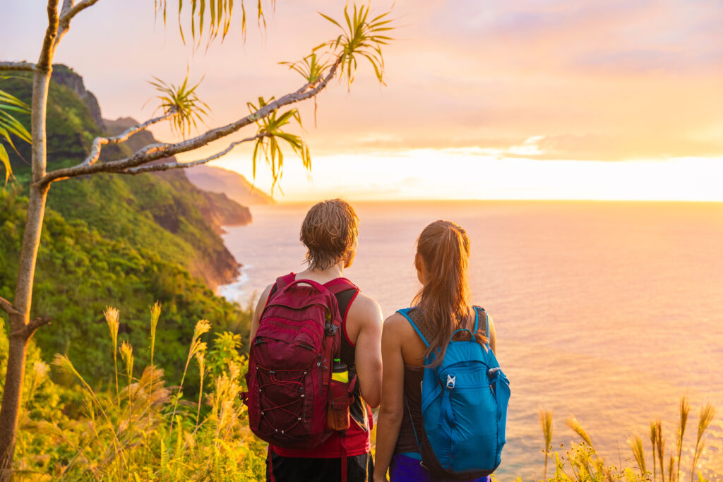 Hawaii hiking hikers hiking on Kalalau trail watching sunset from Na Pali Coast. Tourists couple with backpacks walking outdoor in Kauai island. Summer travel adventure active lifestyle. Romantic getaway. Allegro Luxury Vacations. Brenda Ajay, travel advisor