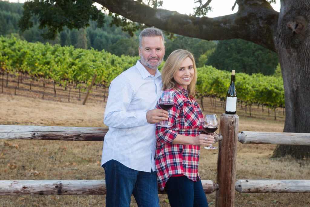 Attractive couple leaning against a fence at a wine vineyard while drinking a glass of wine. Romantic getaway. Napa Valley honeymoon. Allegro Luxury Vacations. Brenda Ajay, Virtuoso Luxury TRavel Advisor