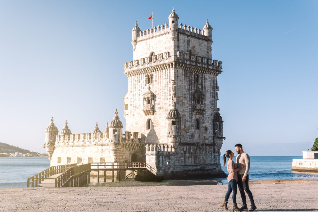 Couple in love embracing and looking at each other in front of the Tower of Belem on a sunny day on the banks of the Tagus River. Romantic getaway. Allegro Luxury Vacations, Brenda Ajay, Virtuoso luxury travel advisor